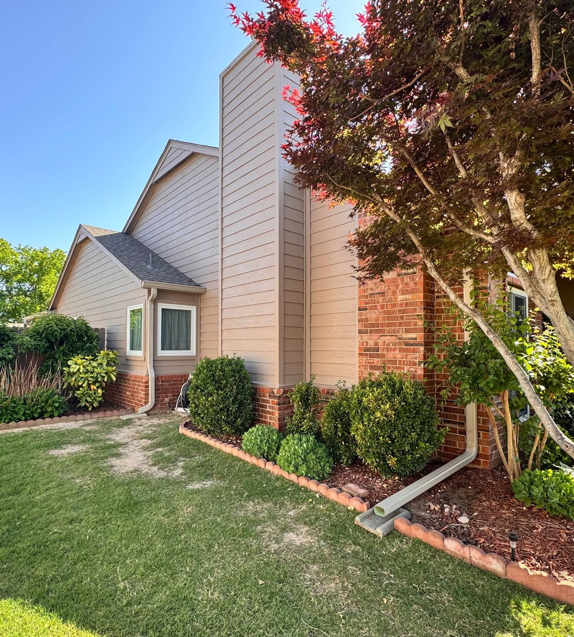 A tan-sided house with a brick base and a Japanese maple tree is shown in a sunny yard.