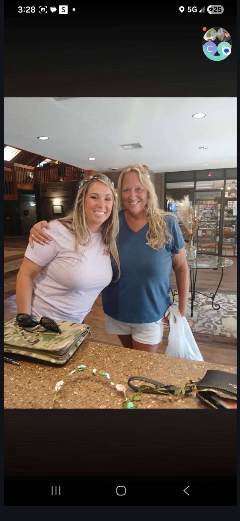 Two smiling women with flower crowns pose for a photo indoors, with a counter and shopping bags visible.