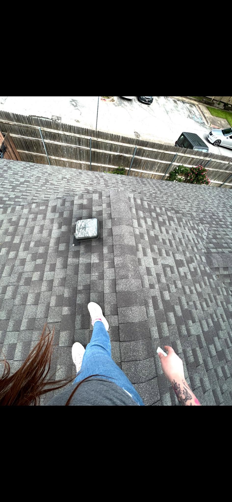 A person stands on a shingled roof, holding a white object, with cars visible beyond a fence.