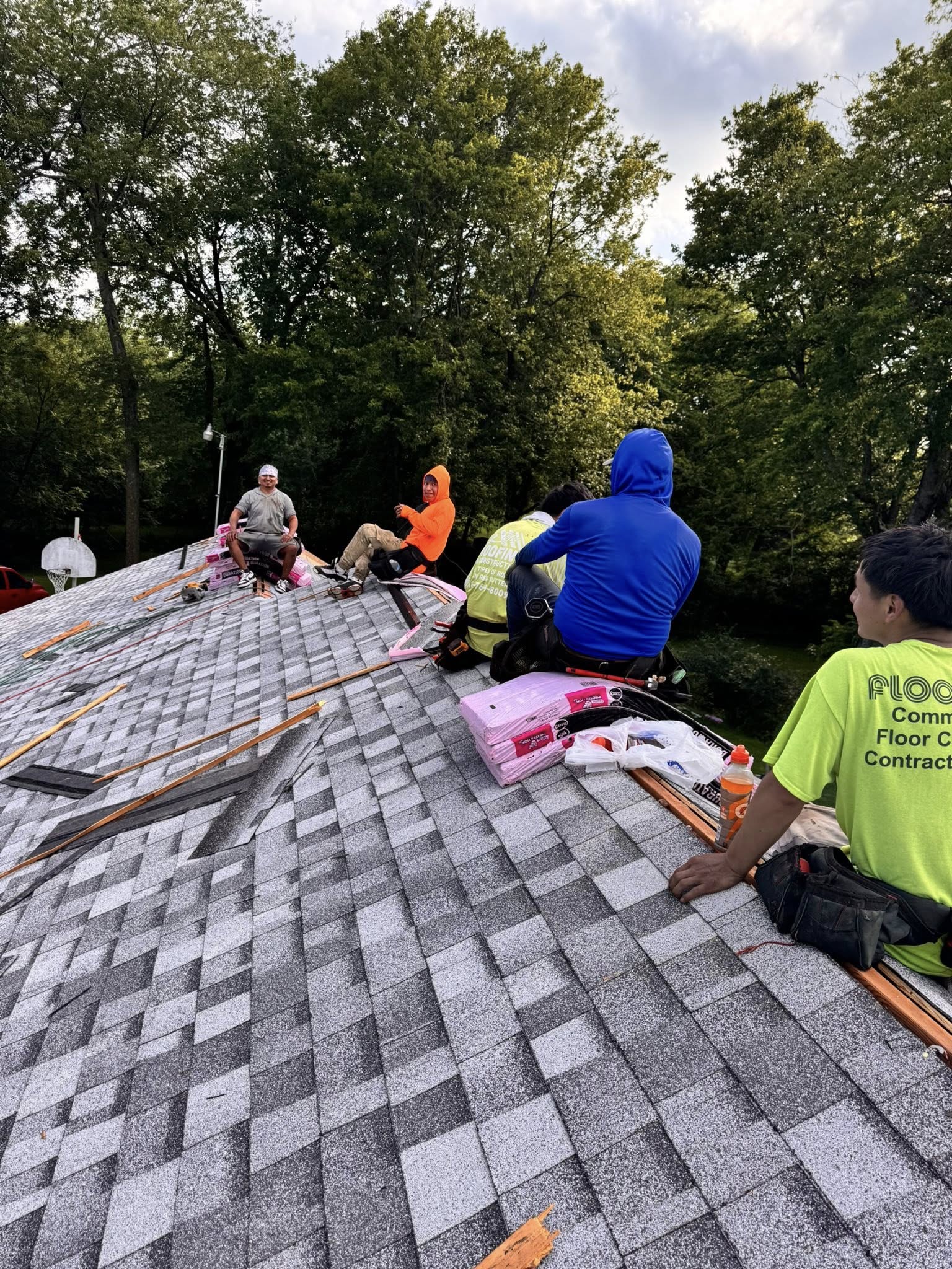 Roofers work on a shingled roof, with tools and materials present, surrounded by trees.