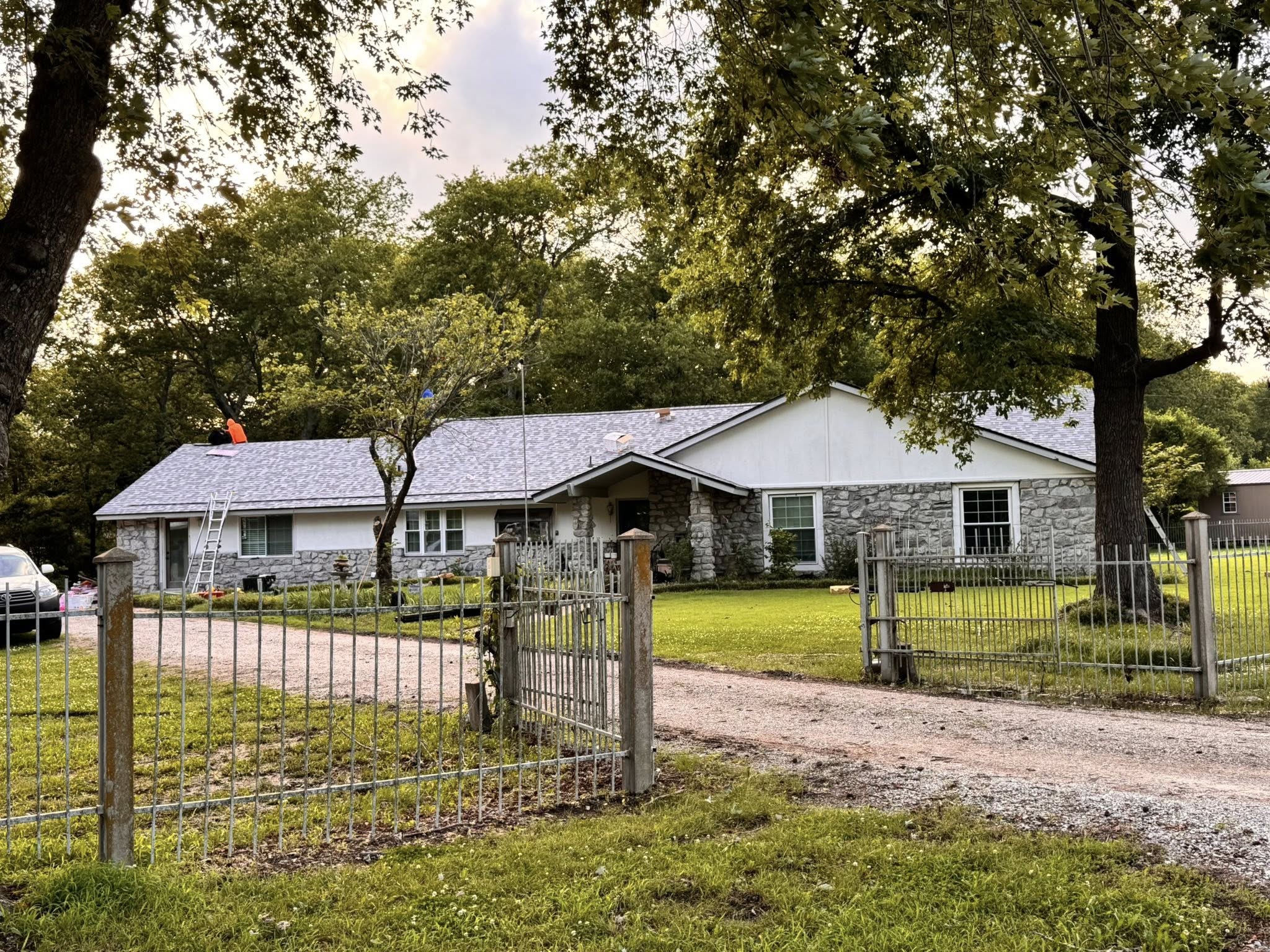 A person in an orange vest works on a house roof, with a fence and trees in the foreground.