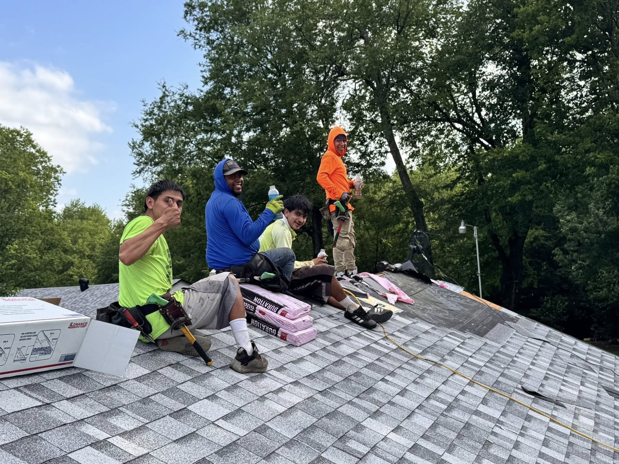 Roofers on a shingled roof with supplies and trees in the background.