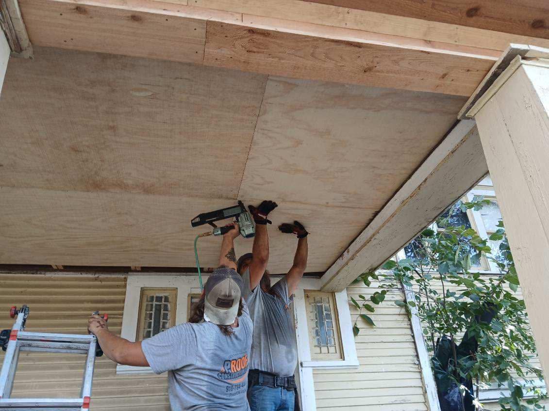 Two men are working on a porch ceiling, using a nail gun to attach plywood.