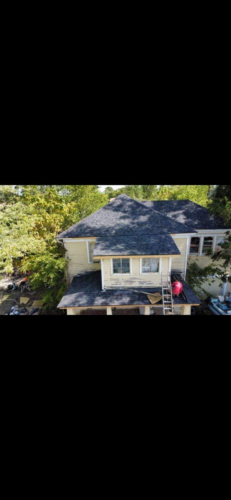 A person in a red shirt works on a roof of a yellow house surrounded by trees.