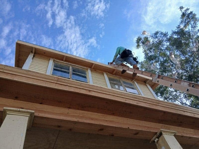 A construction worker on a ladder works on the roof of a yellow house under a blue sky.