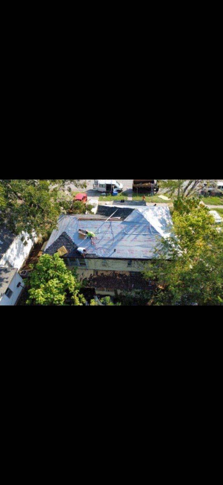 Workers tarp a damaged roof on a house surrounded by trees and vehicles.