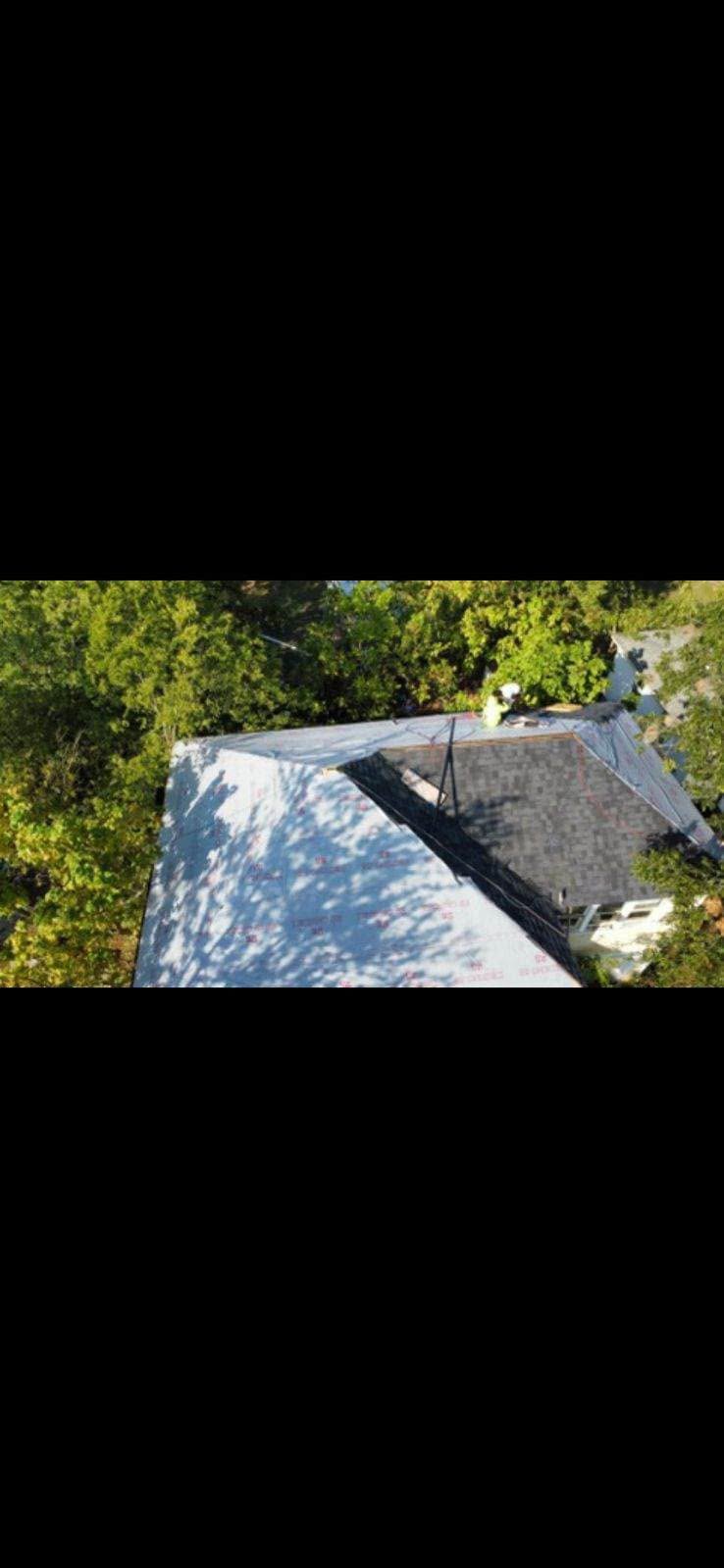 A person works on a partially shingled roof of a house surrounded by trees.