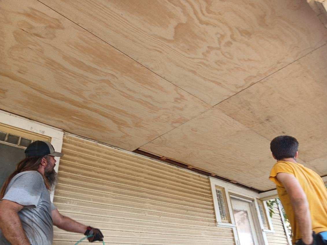 Two men work on a plywood ceiling on a porch.