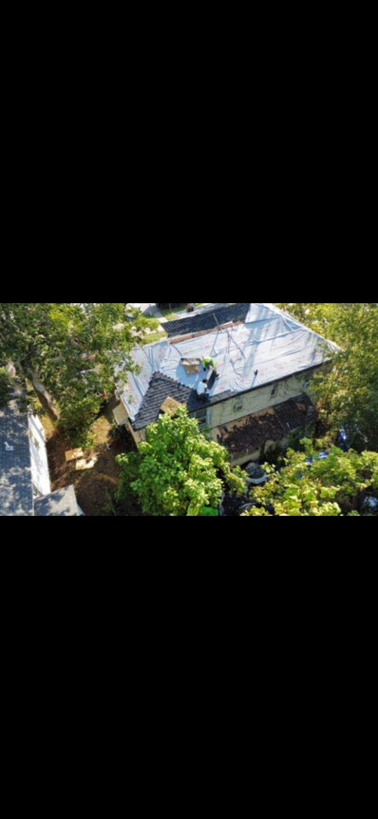 Workers are on a tarp-covered roof of a house surrounded by trees.