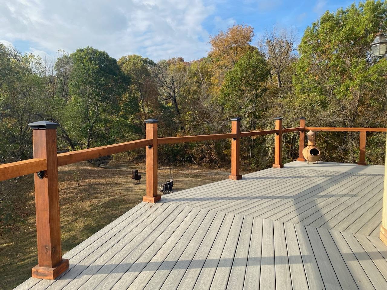 A wooden deck with railings overlooks a wooded area with a chiminea on a sunny day.
