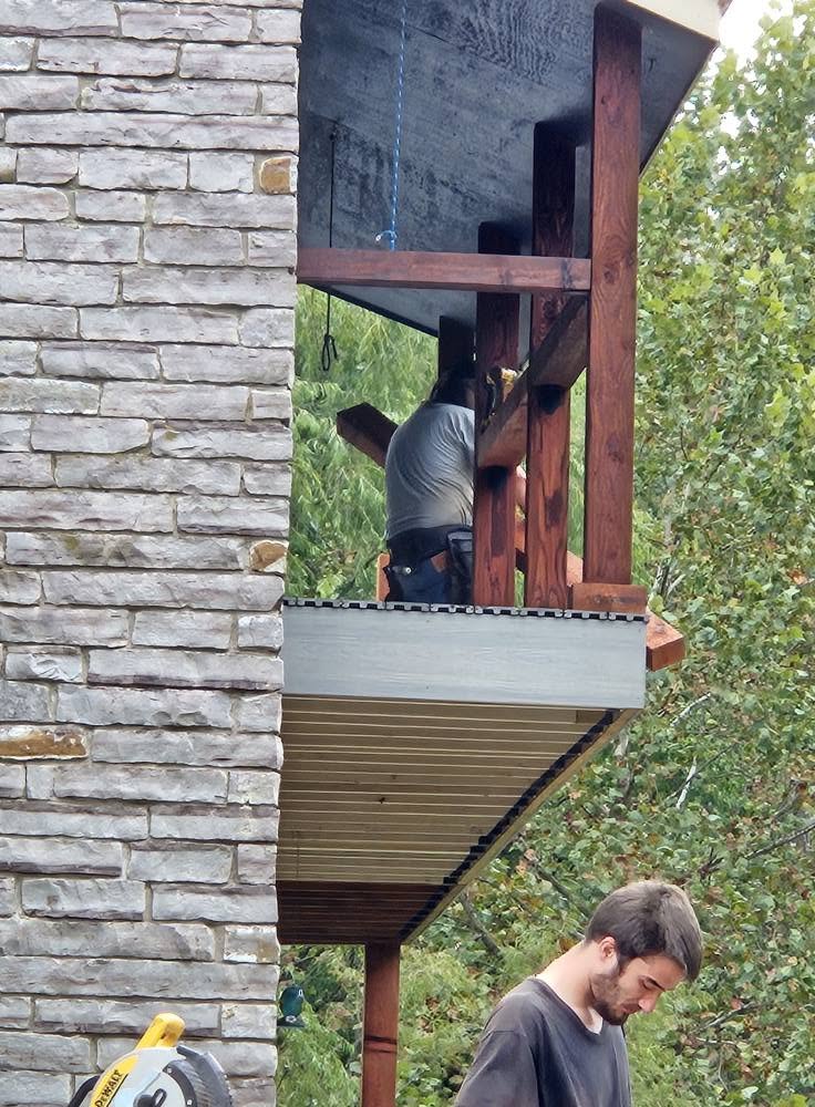 Two men work on a wooden structure next to a stone wall, with trees in the background.