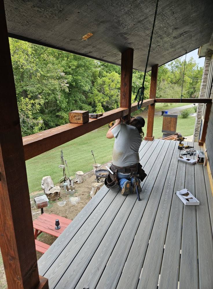 A construction worker installs a deck railing on a porch overlooking a wooded area.