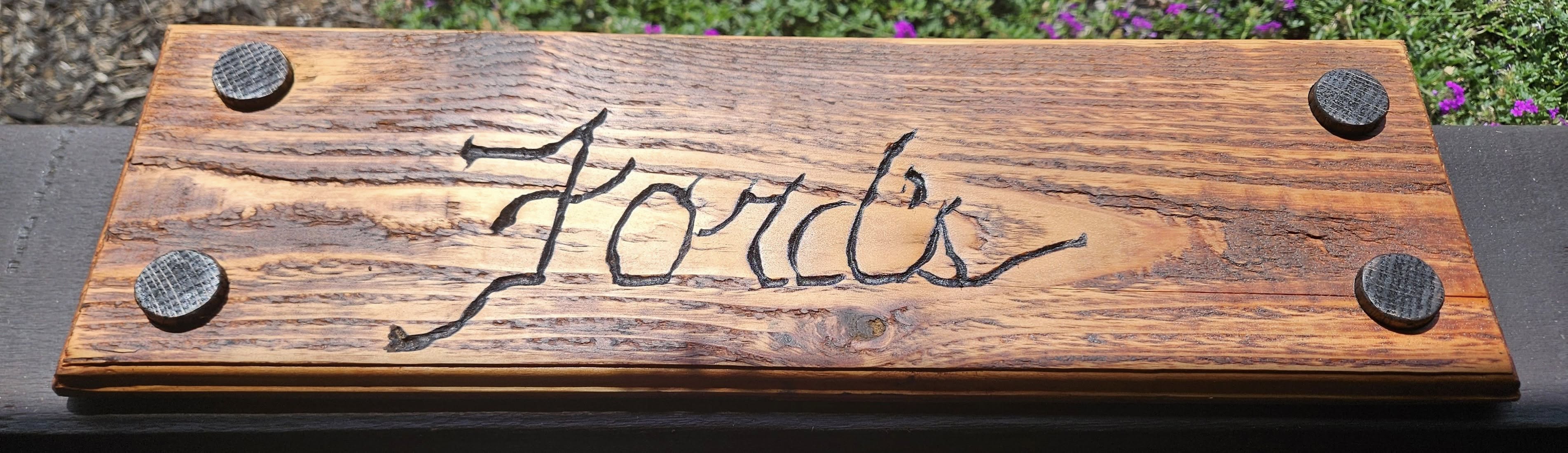 A wooden sign with "Ford's" carved into it, mounted with four circular fasteners.
