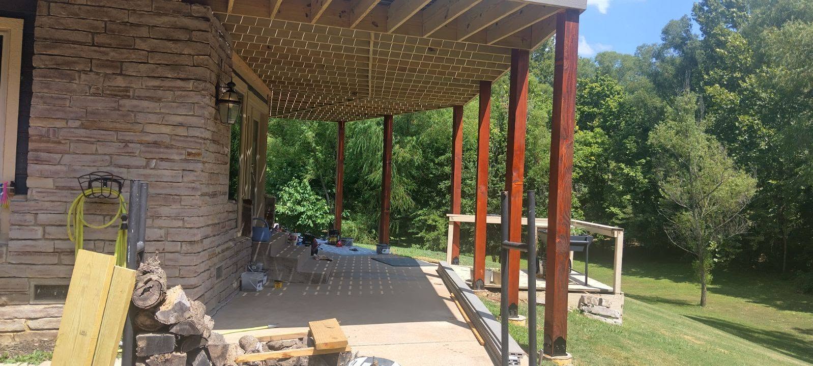 A covered porch with wooden beams and stone siding is shown in a wooded area.