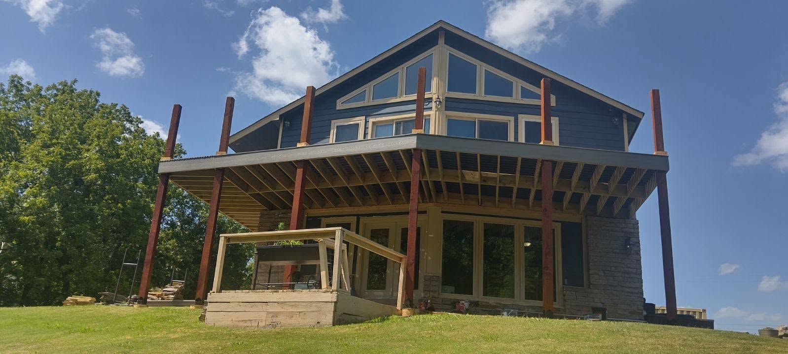 A blue house with a large deck under construction on a grassy hill.