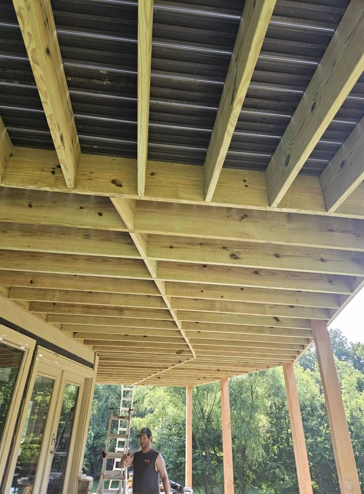 A man stands under a new wooden deck structure with a ladder nearby.