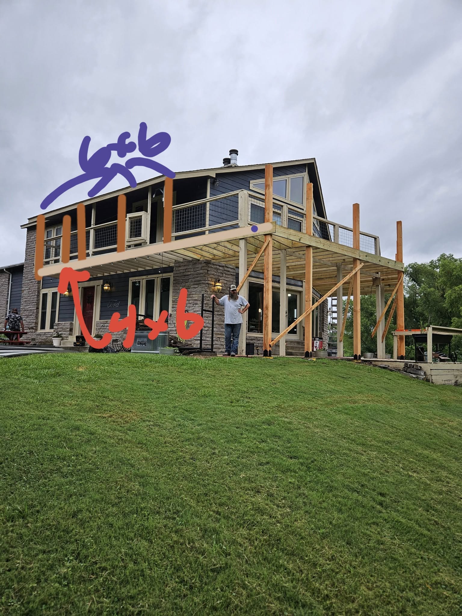 A man stands on a grassy hill near a house with a new deck under construction.