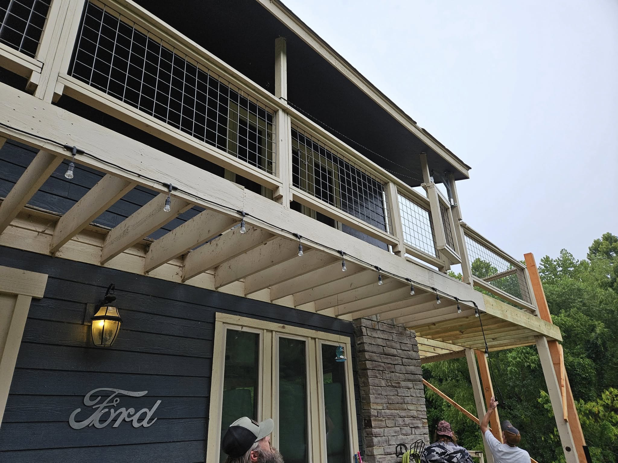 Two people are working on a wooden deck with string lights attached to the underside.
