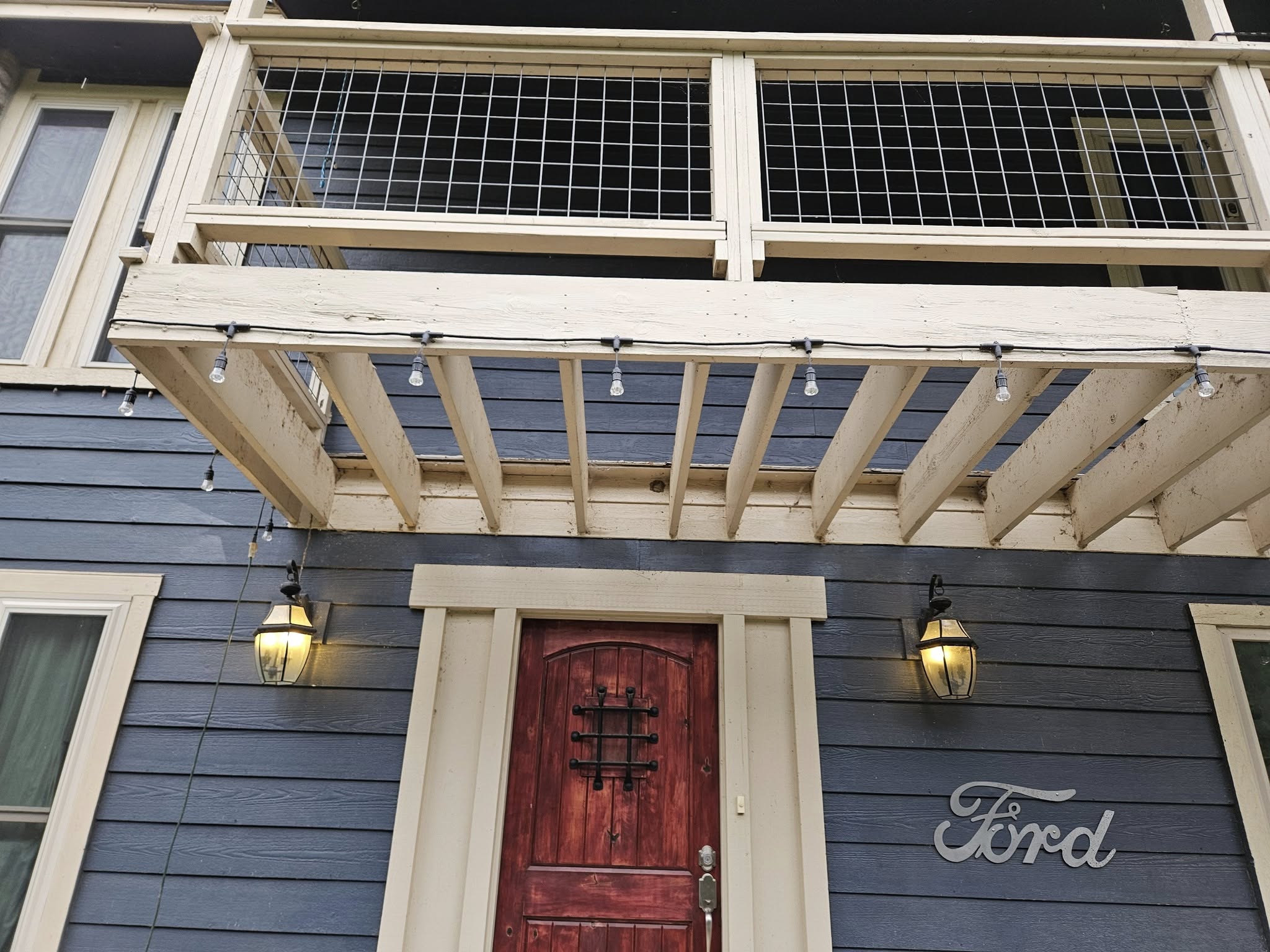 A rustic wooden door with a metal grate is framed by a pergola with string lights, on a blue-sided house.