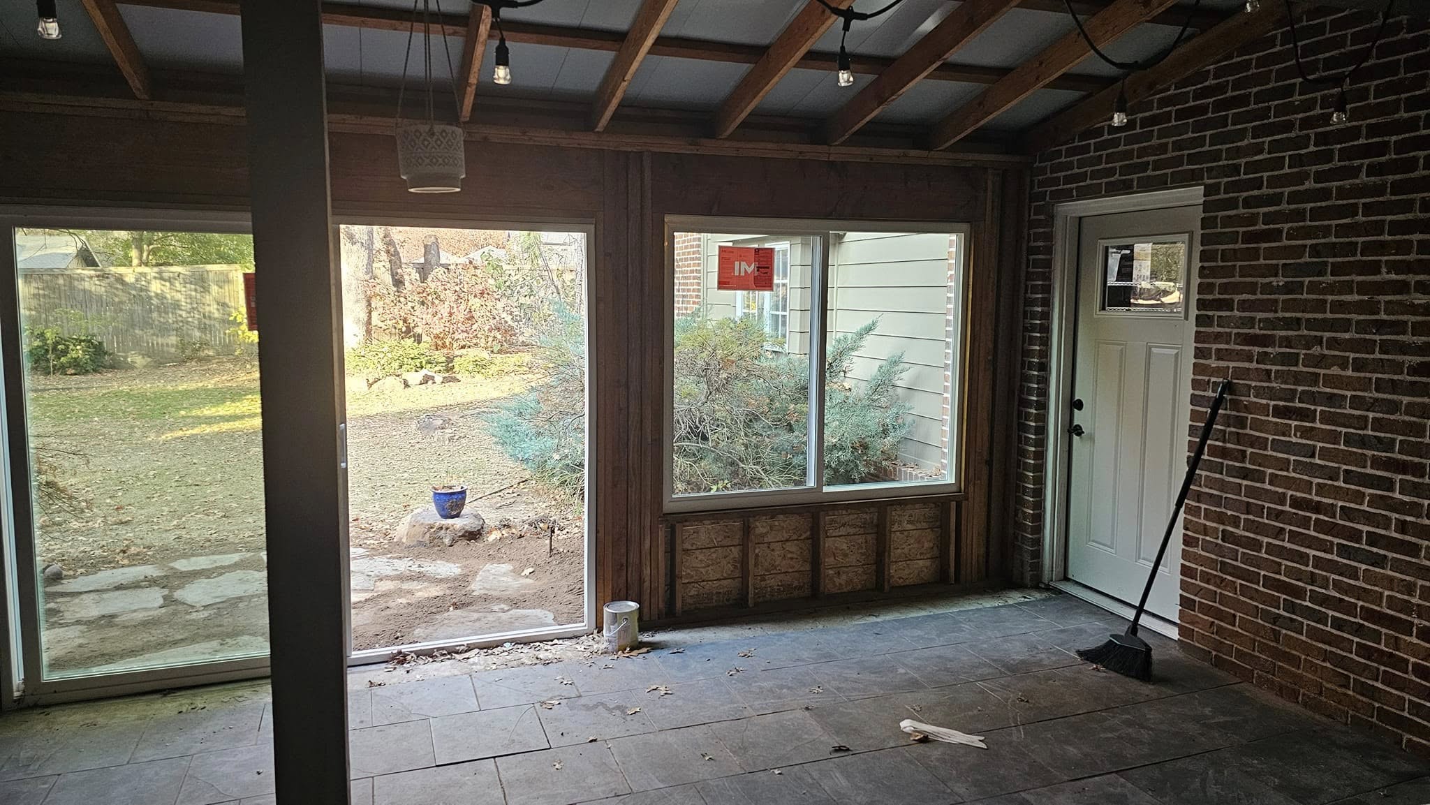 An unfinished sunroom with large windows, a door, and a broom on a brick wall.