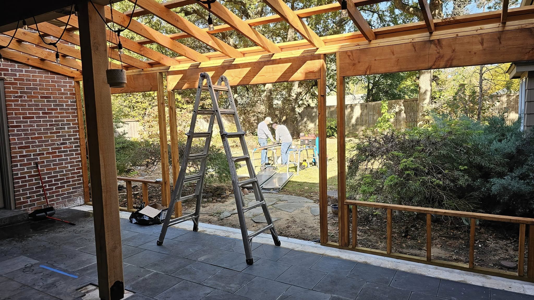 Two men work on a wooden pergola construction in a backyard with a ladder and tools.